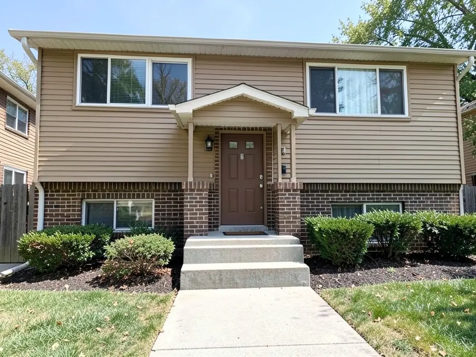 Split-level style house with tan vinyl siding above brown brick, a small gabled entry roof, a brown front door and three plain grey concrete steps down to a concrete walkway