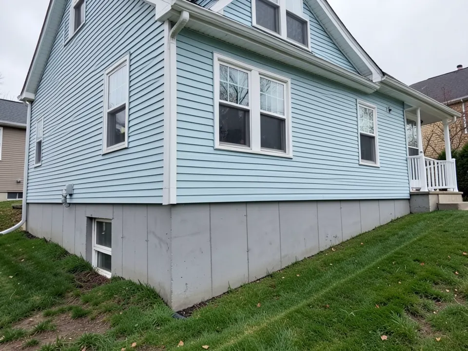 AI-drawn stand-in of a pale blue two-story house on a sloping lawn, with a tall band of bare grey poured concrete foundation and a basement window showing below the siding
