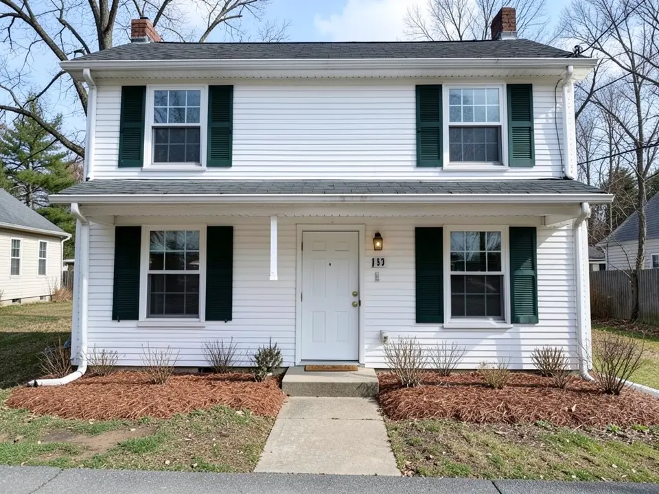 Two-story colonial house with white vinyl siding, dark green shutters, a plain white front door and bare mulch beds