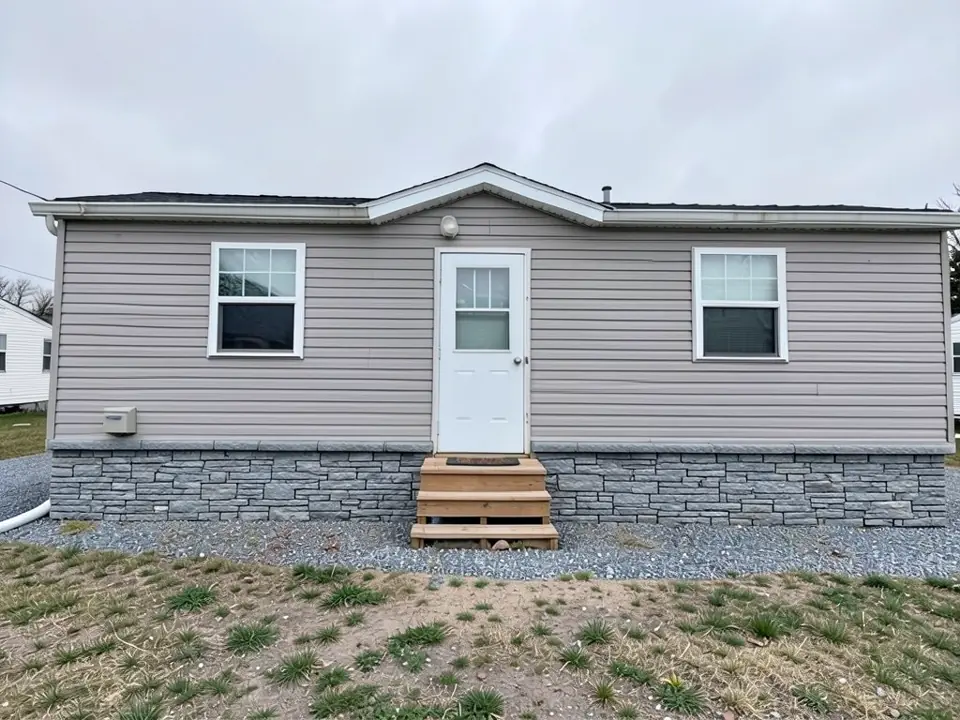 The same single-wide with grey stacked-stone look skirting panels running the full length of the base; siding, windows, door and wooden steps unchanged