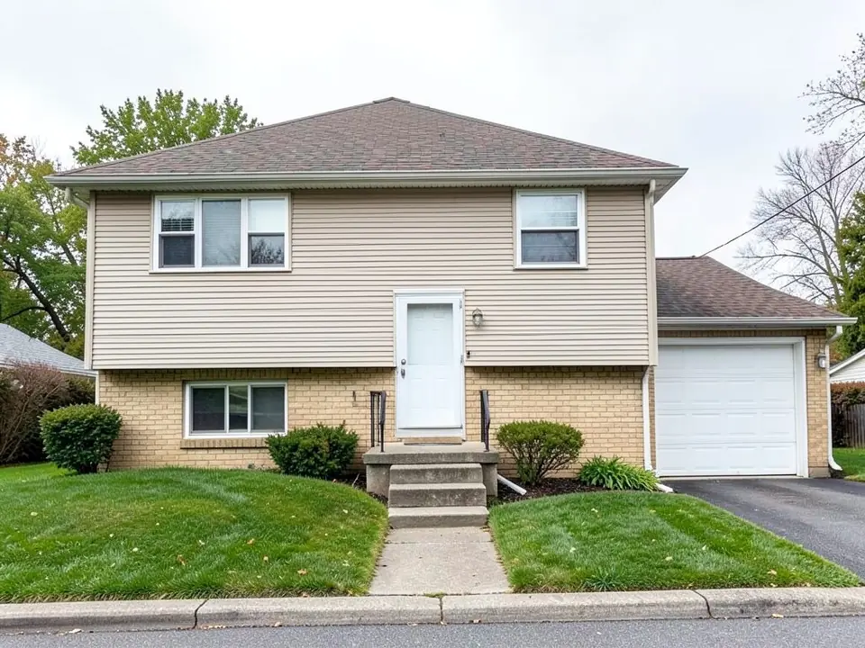 Raised ranch (bi-level, split-entry) house with a tan brick lower floor, beige vinyl siding above and a plain white front door at mid-height up concrete steps