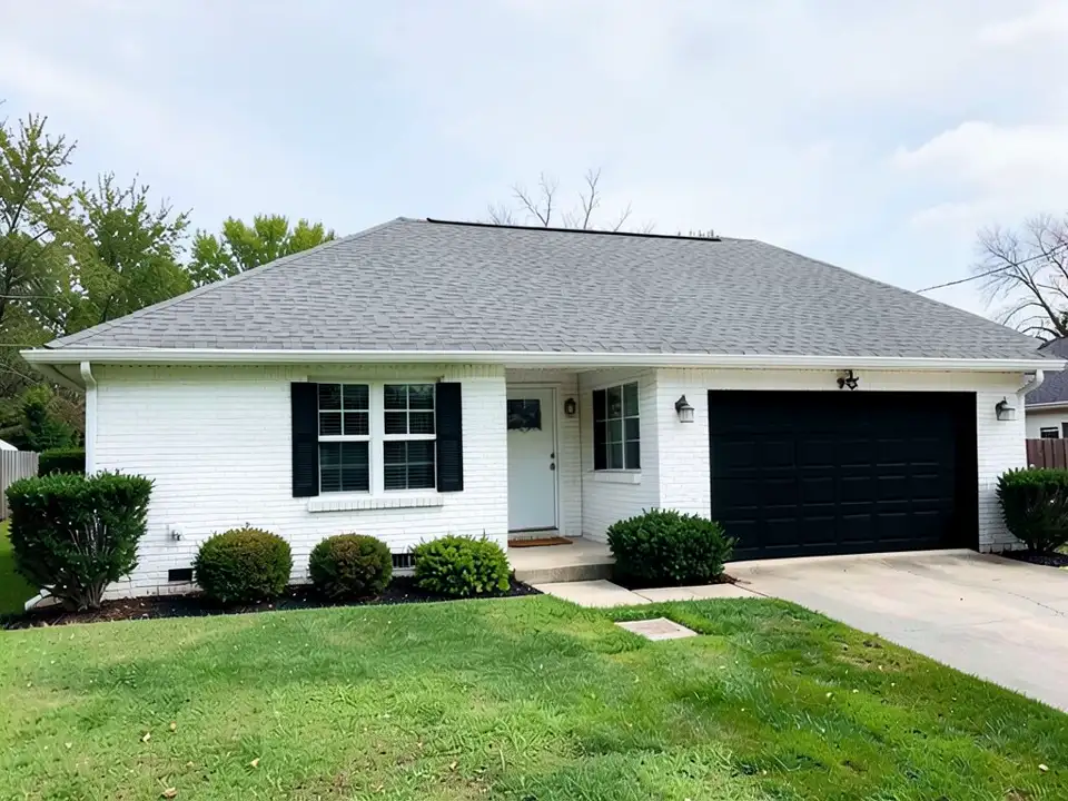 The same house with a black garage door and black shutters on the left window; the white brick, white trim, roof, driveway, shrubs and lawn kept